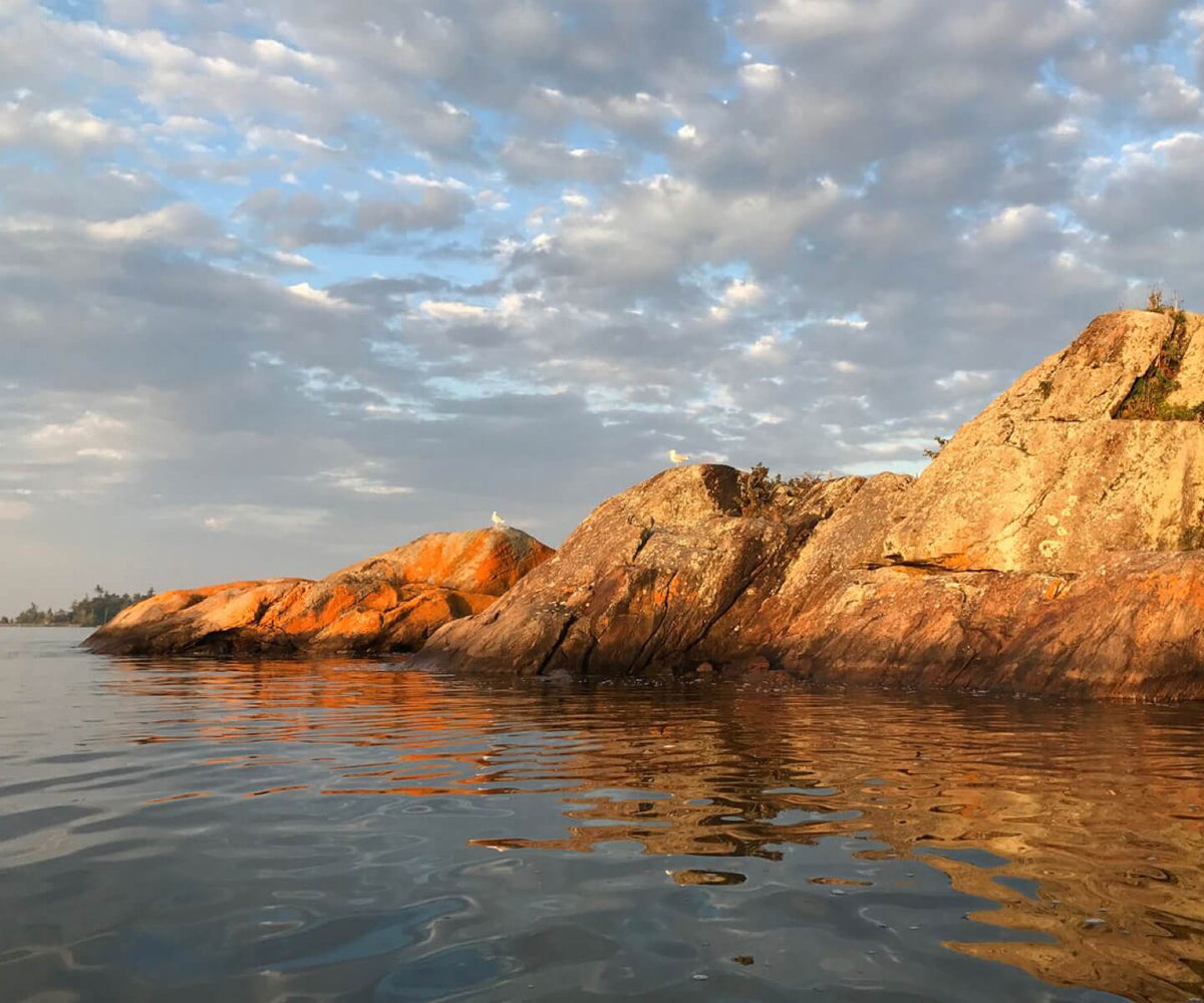 Rocky shoreline with large tan and orange-brown cliffs reflected in calm water under a cloudy blue sky.